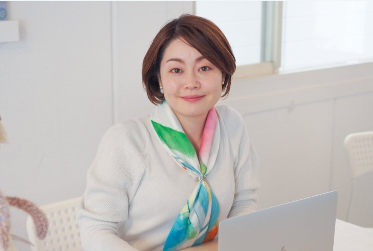 白い部屋でスカーフを巻き、ノートPCの前に座って微笑む女性