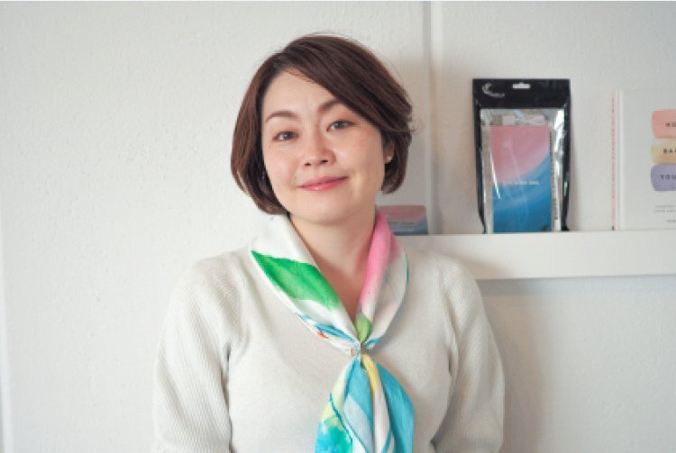 白い棚の前に立ち、スカーフを巻いて微笑む女性