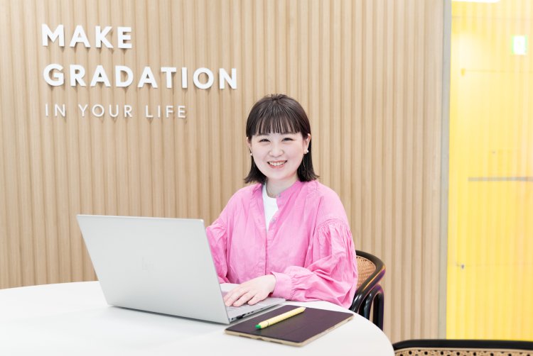 GRADATION IN YOUR LIFEのロゴが並ぶ店内でノートPCを開き、笑顔でこちらを見るkahoさん