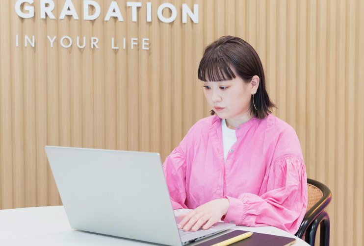 GRADATION IN YOUR LIFEの看板前でノートPCに向かうkahoさんの写真