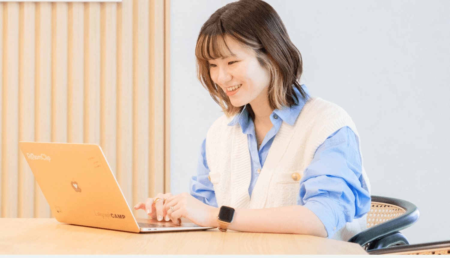 木目調のオフィスでオレンジ色のノートパソコンに向かい微笑む彩華さん