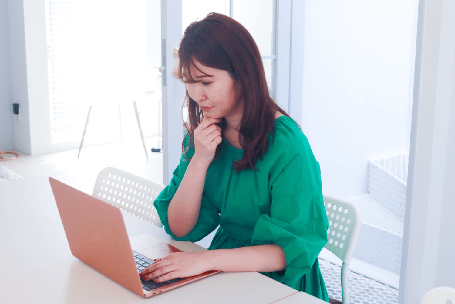 白い室内でノートPCに向かい、考えるしぐさで作業する緑ブラウスの女性