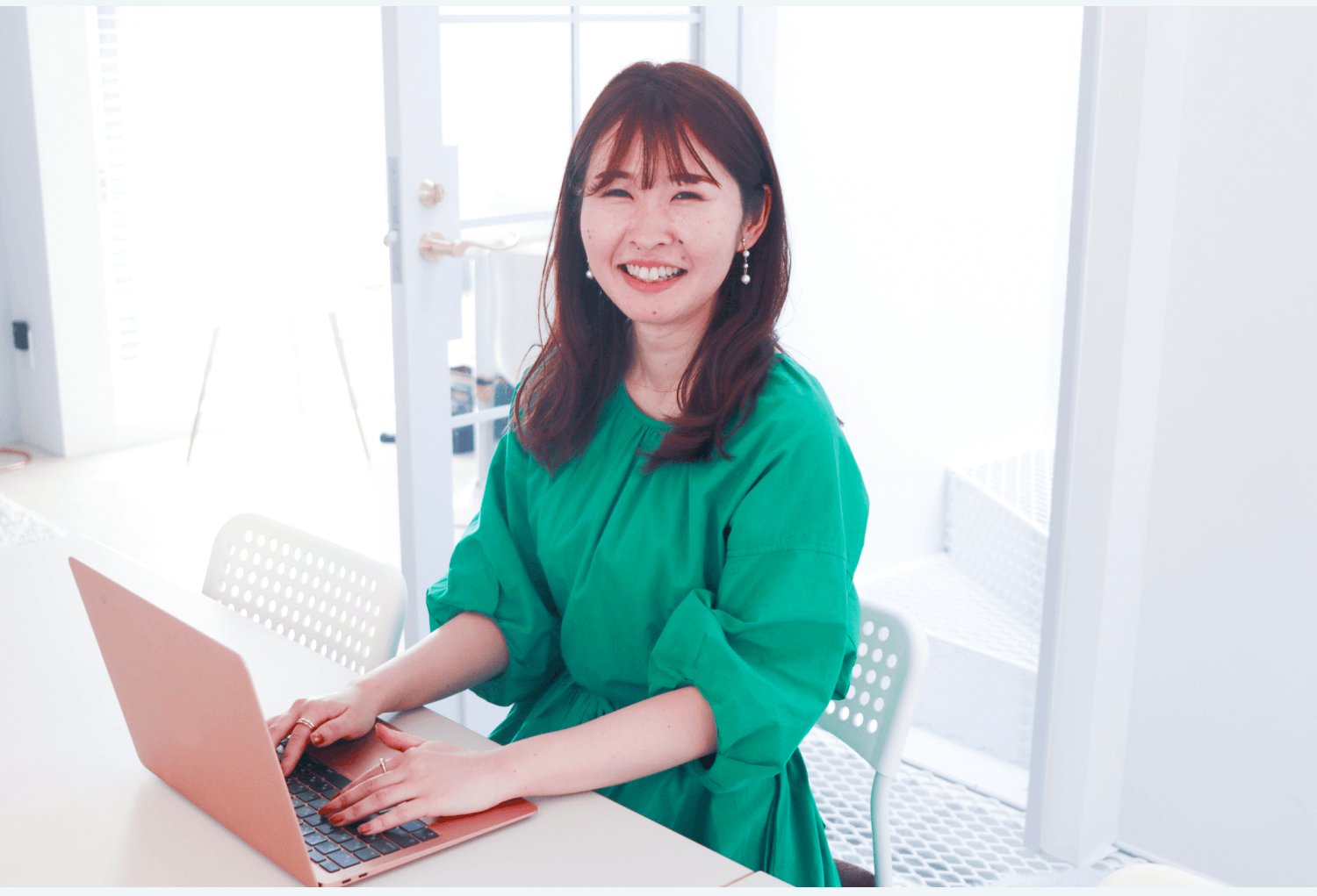 白い室内でノートPCに向かい微笑む緑ブラウスの女性