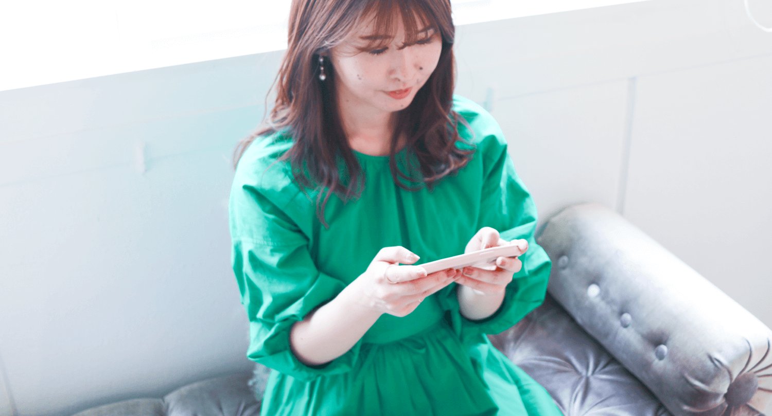グレーのソファに座り、スマートフォンを操作する緑ブラウスの女性