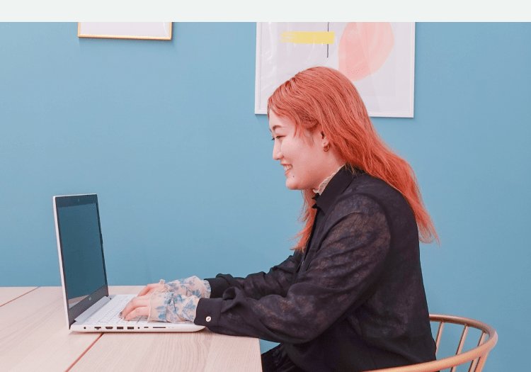 青い壁を背景に、木製デスクでノートPCを操作しながら微笑む女性の横顔