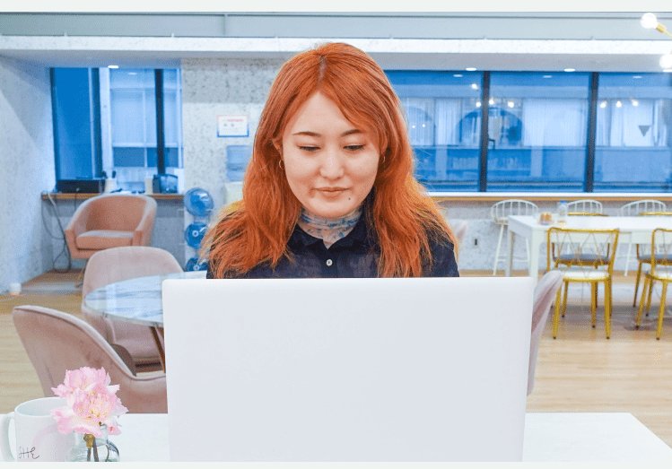 カフェ風のスペースでノートPCに向かう女性。花瓶に活けたピンクの花とマグカップが手前にある