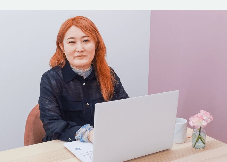 白とピンクの壁を背に、ノートPCに向かい正面を見て微笑む女性。デスクに花瓶とマグカップ