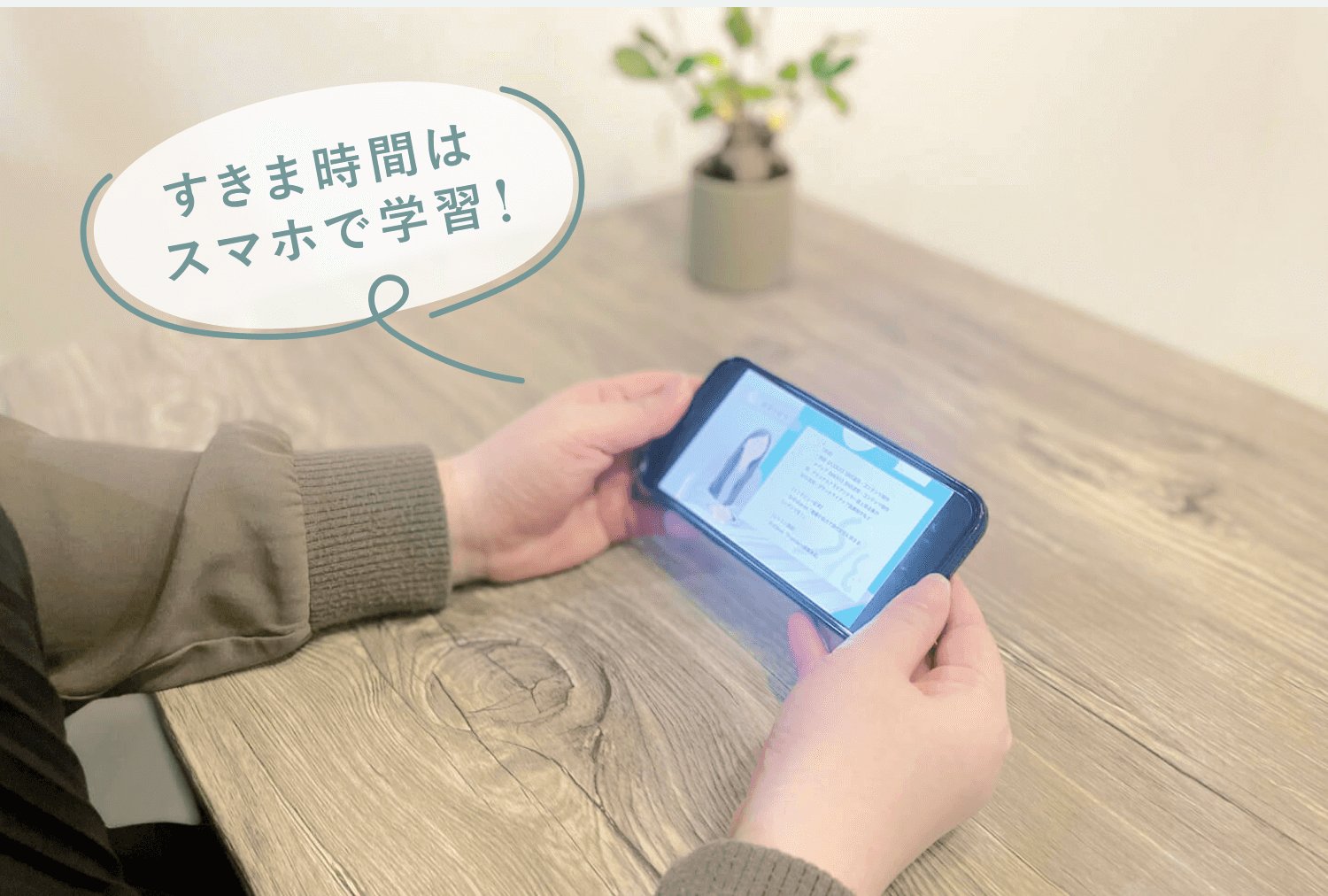 木製テーブルの上でスマートフォンを操作する手元。「すきま時間はスマホで学習！」という吹き出しキャプション付き