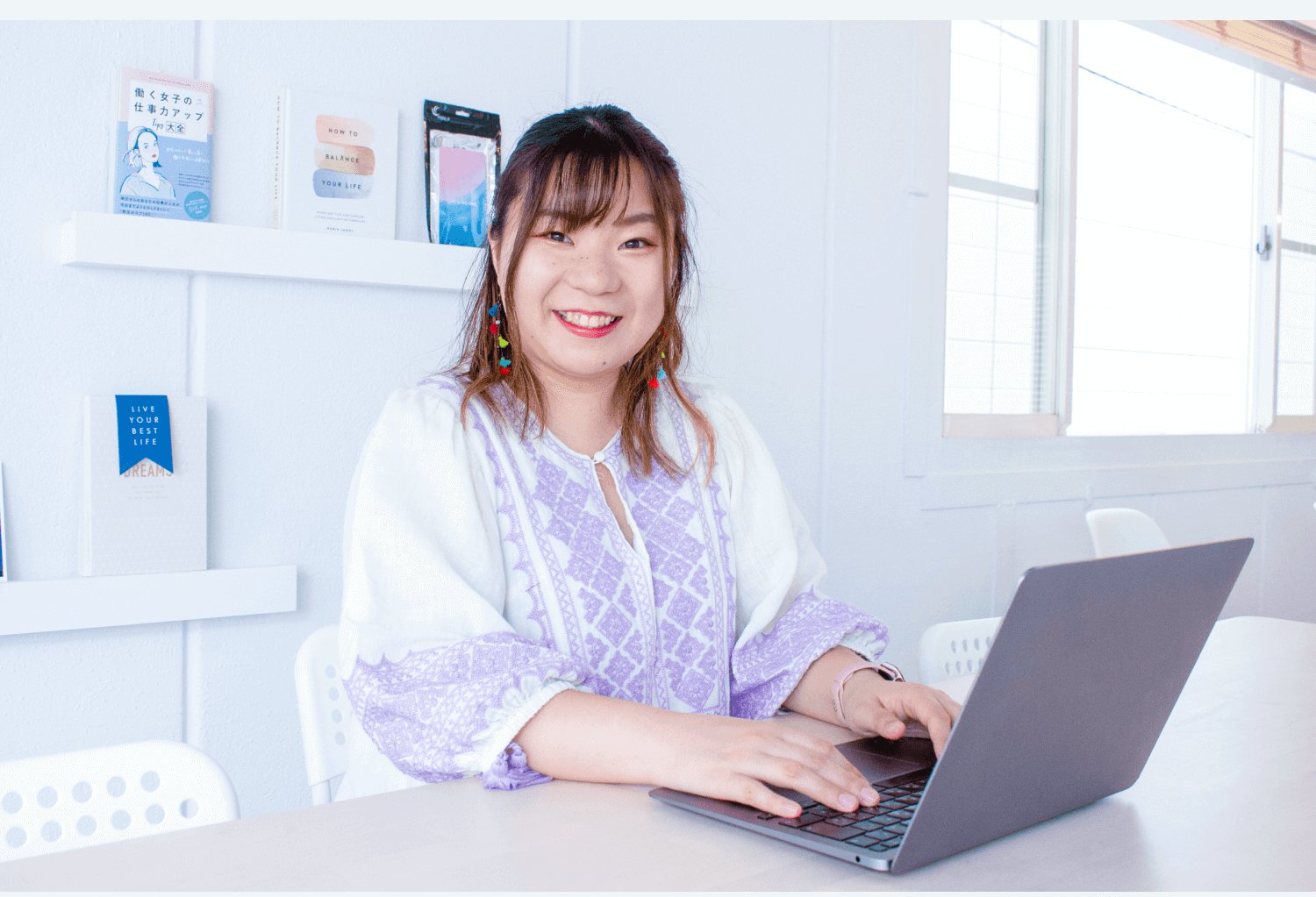 ノートパソコンで作業する本人。背景の棚に本や「LIVE YOUR BEST LIFE」のリボン型オブジェが並ぶオフィスの一室