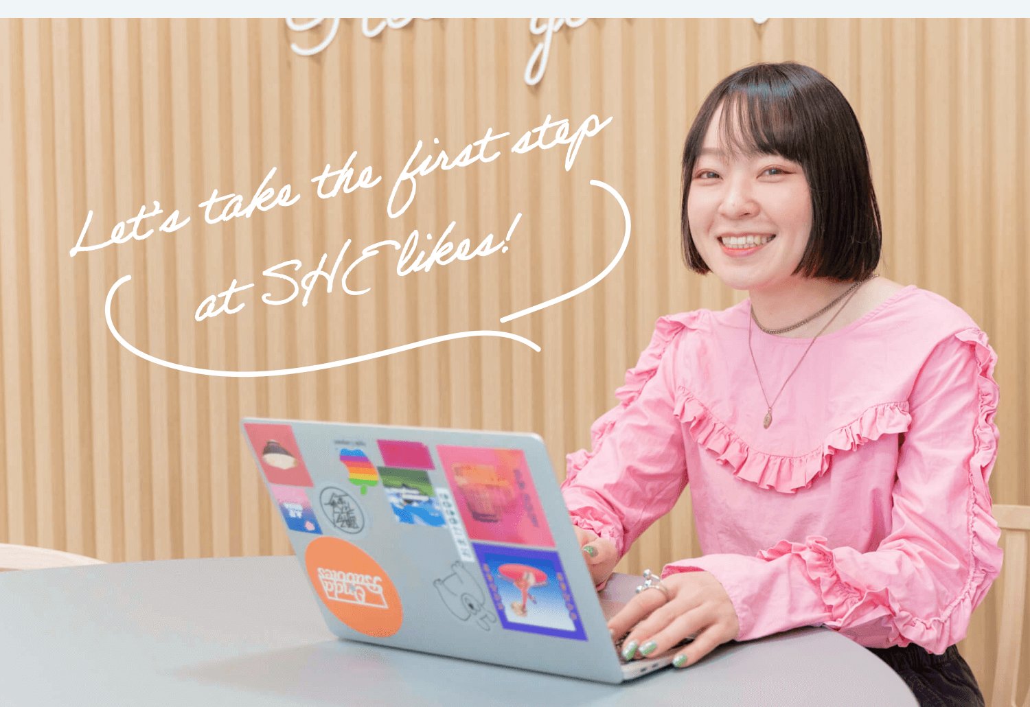木目パネルの壁を背景に、ステッカーを貼ったノートPCを操作しながら微笑む本人（ピンクブラウス）。背景に手書き風の文字装飾「Let's take the first step at SHElikes!」
