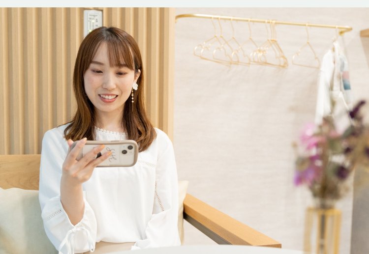 スマートフォンを手に微笑む女性。背景にハンガーラックと花