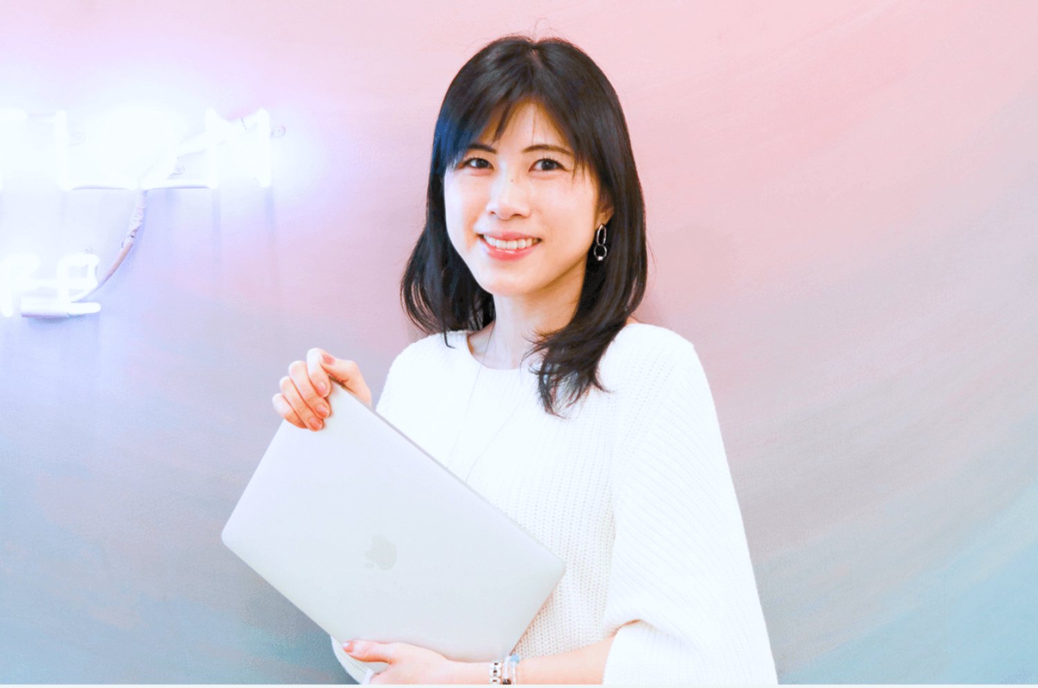 ピンク〜紫のグラデーション壁の前で白いノートPCを抱えて微笑むsatominさん