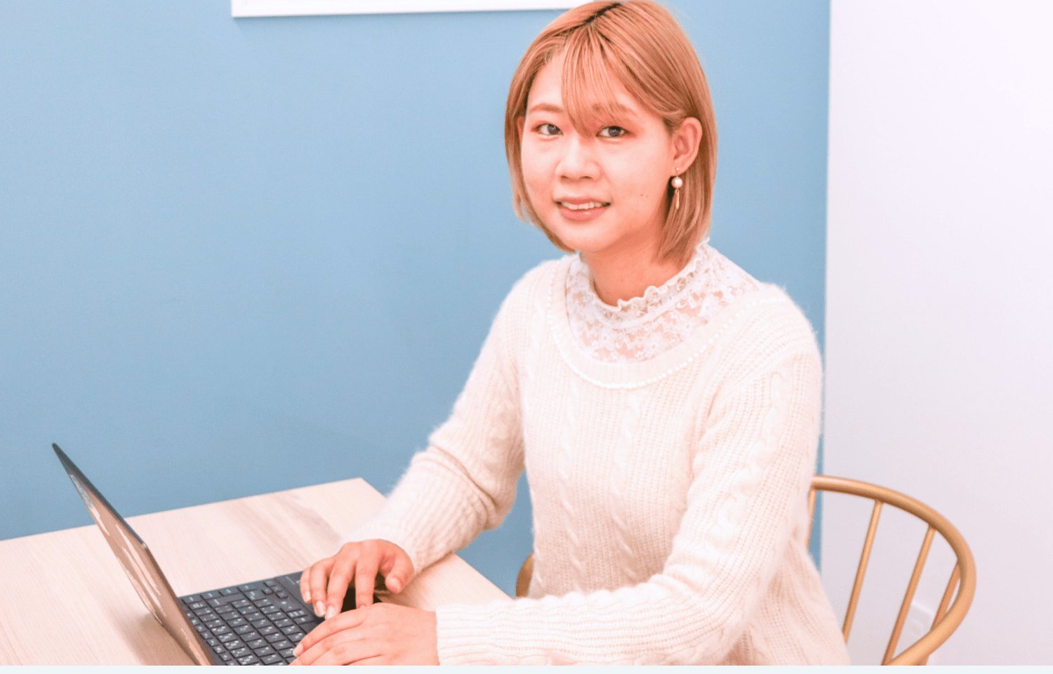 青い壁の前でノートパソコンを操作し正面を見て微笑むちさとさん