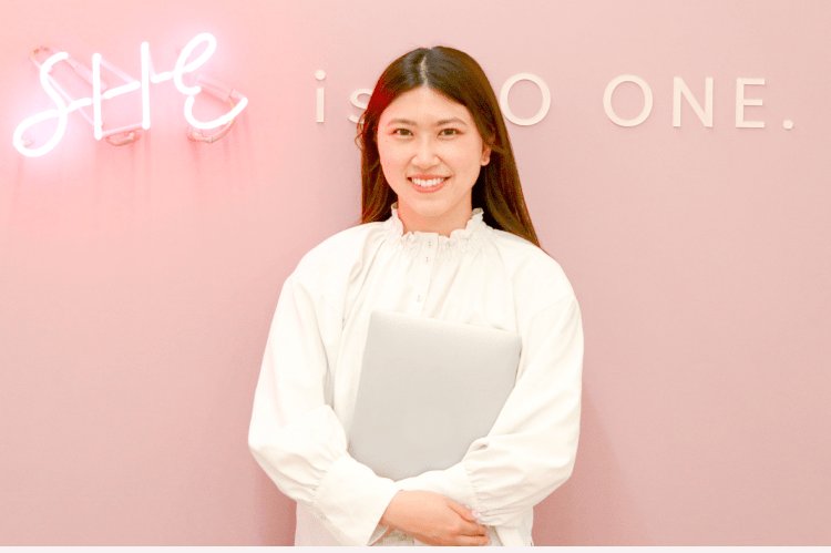 ピンクの壁にある「SHE is NO ONE.」のネオンサインの前で、ノートPCを抱えて微笑むしおりさん