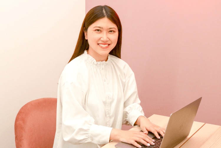 ピンクの壁の前でノートPCを操作しながら微笑むしおりさん