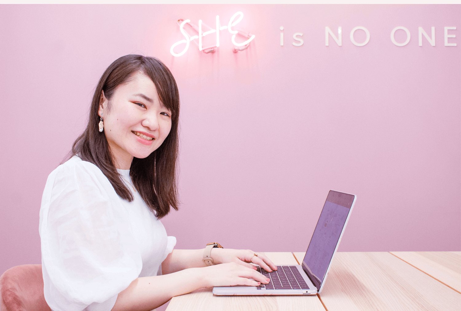 ピンクのネオンサイン「SHE is NO ONE.」の壁の前でノートPCを操作するぺちさんの上半身写真