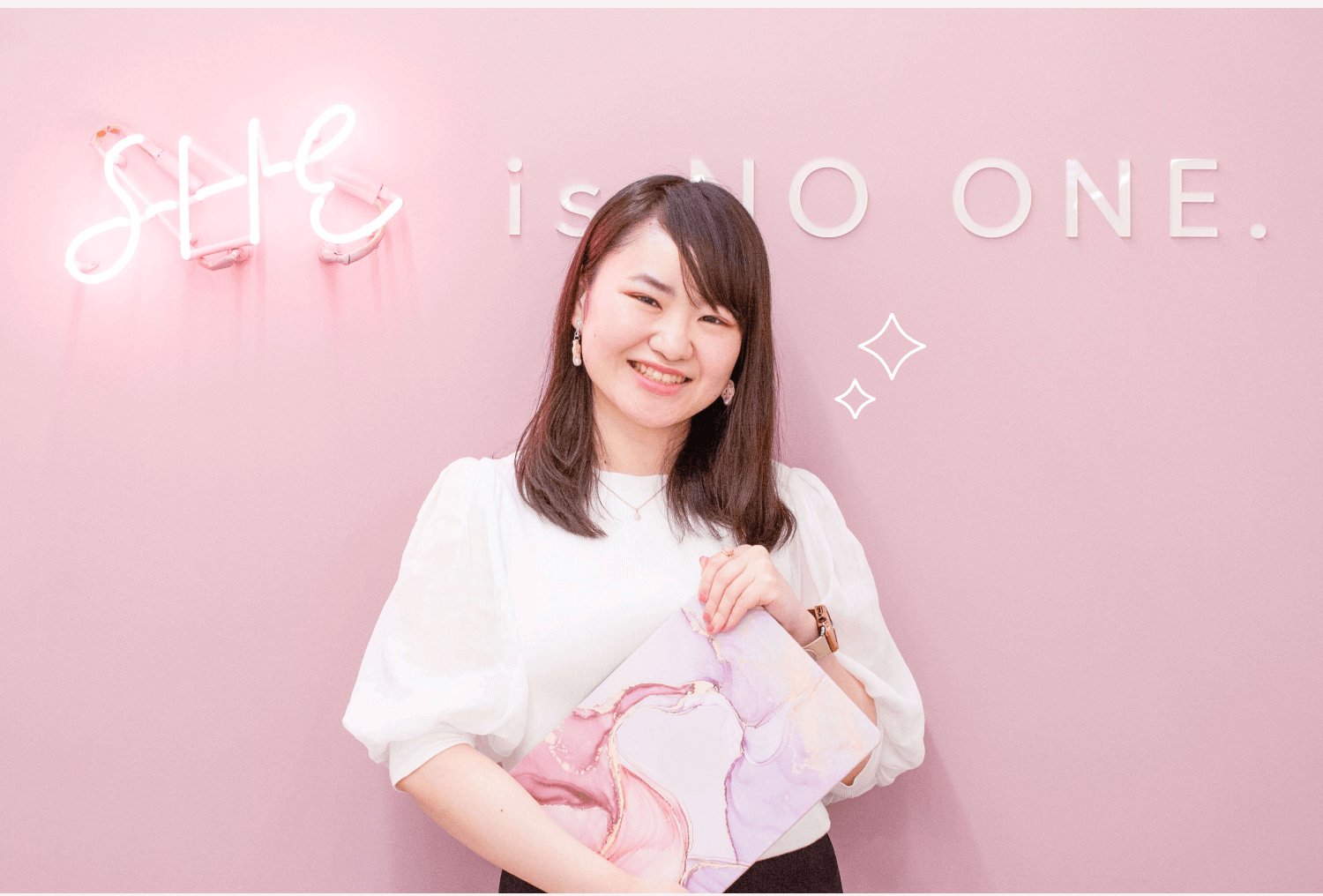 ピンクのネオンサイン「SHE is NO ONE.」の壁の前で水引アクセサリーの水彩柄フォルダーを抱えて微笑むぺちさんの上半身写真
