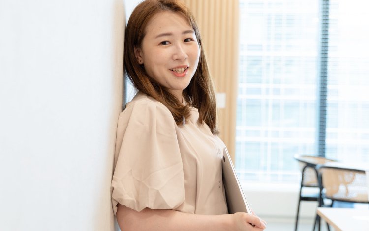 窓辺の壁に寄りかかりファイルを抱えて微笑むゆうきさん