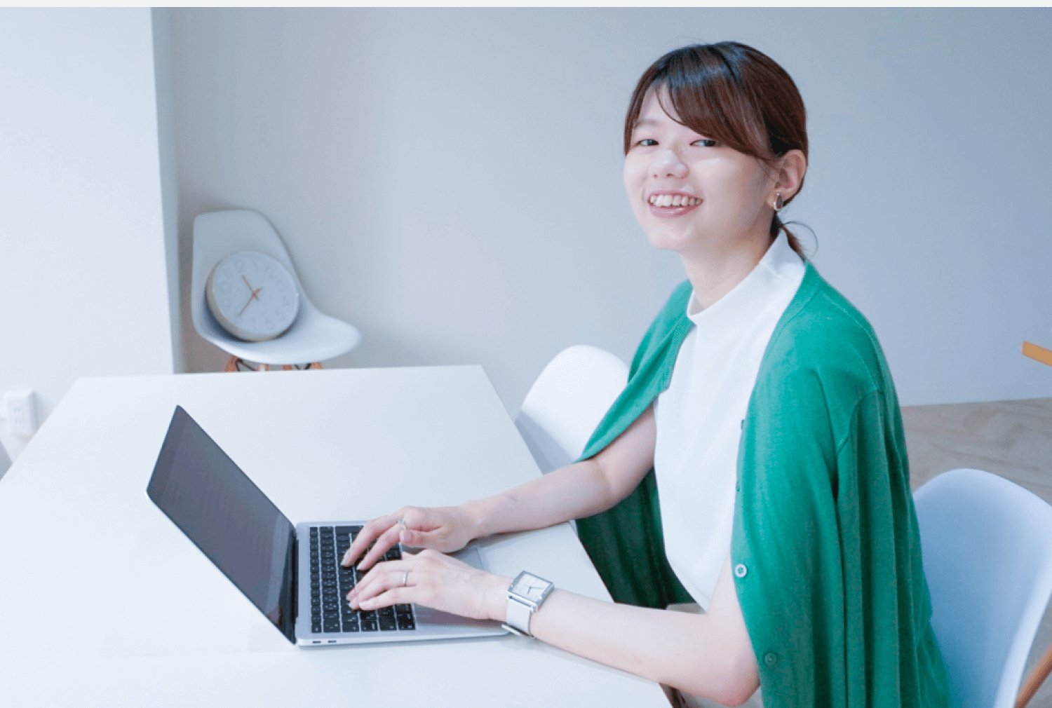 ノートパソコンに向かい、振り返って微笑むほたてさん。白い部屋のテーブルで、背景に椅子と時計が見える