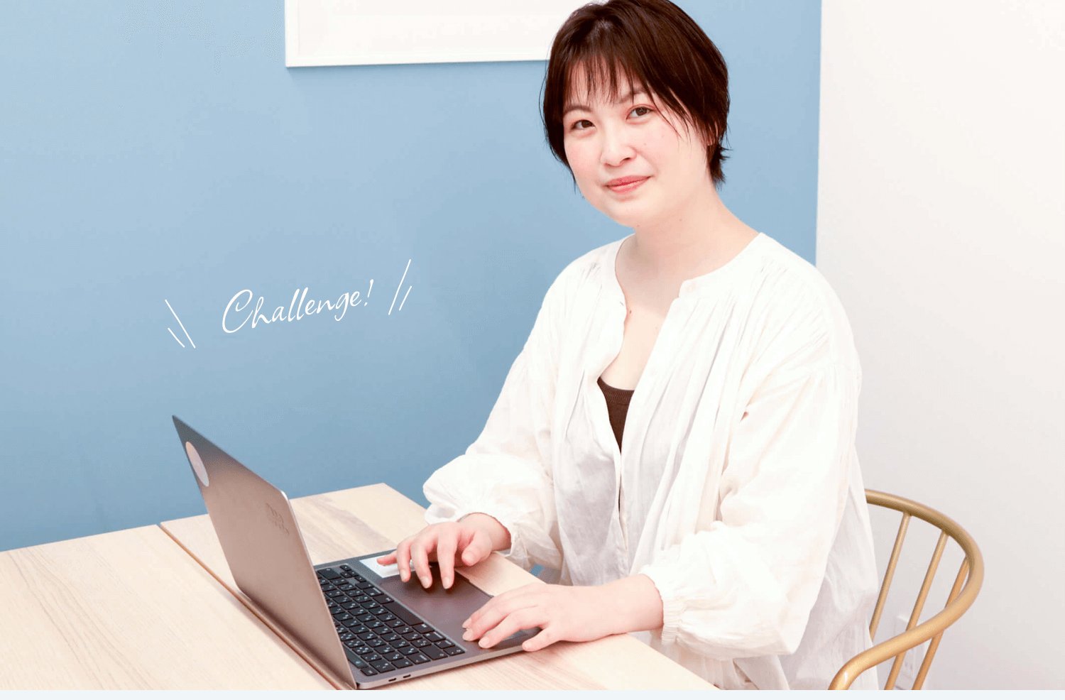 木製テーブルでノートPCを操作しながらこちらを見る女性。壁に「Challenge!」の手書き風装飾文字