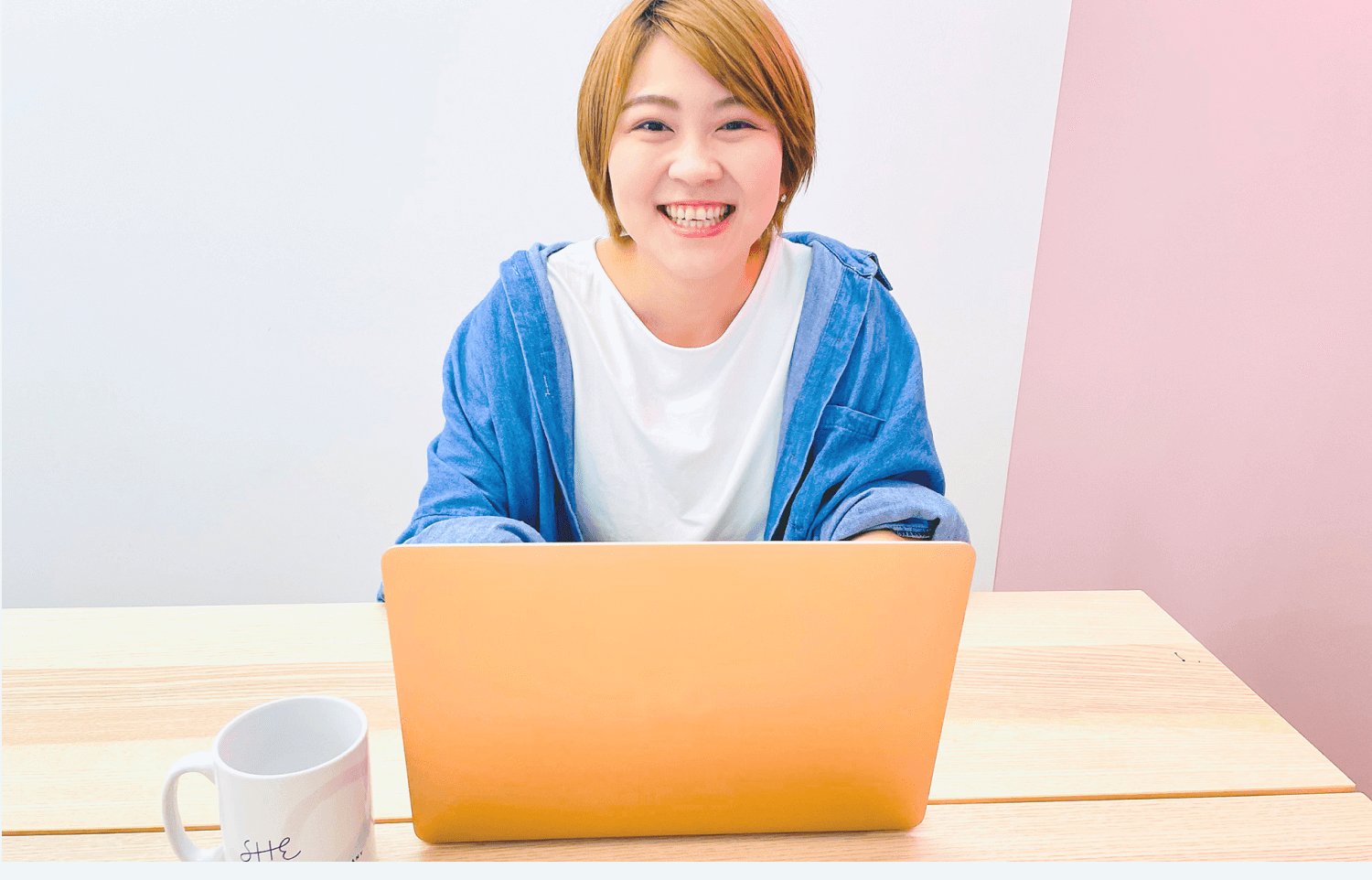 ピンク色の壁を背景に、オレンジ色のノートパソコンに向かい微笑むなりーさん