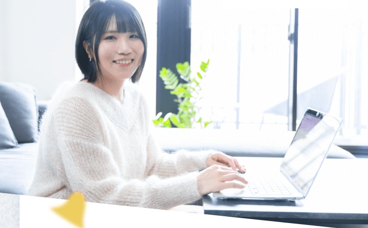 窓辺のテーブルでノートPCを操作しながら微笑むTomoyoさんの写真