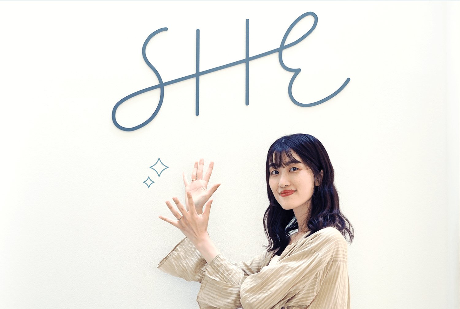 SHEのロゴサインの前で微笑みながら手を挙げるかえさん