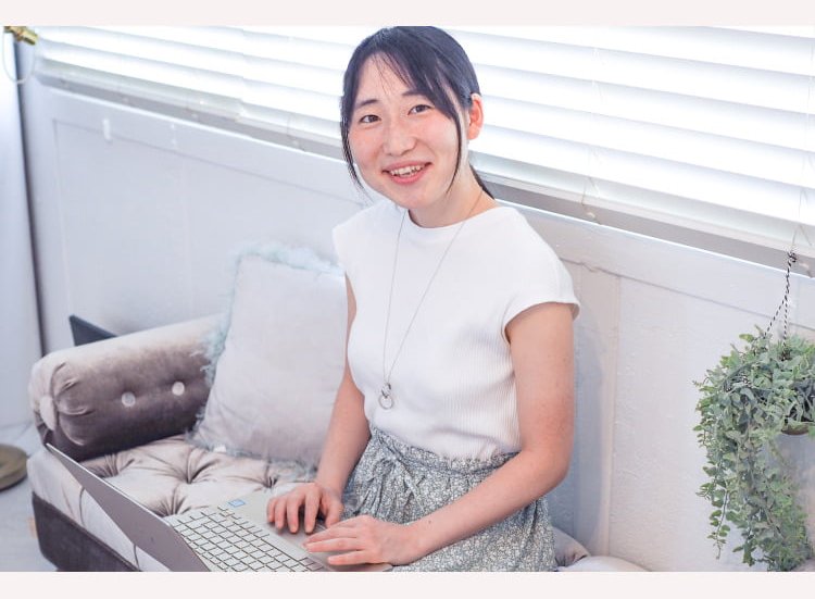 窓辺のソファに座り、ノートPCで作業しながら微笑むこじゆかさん
