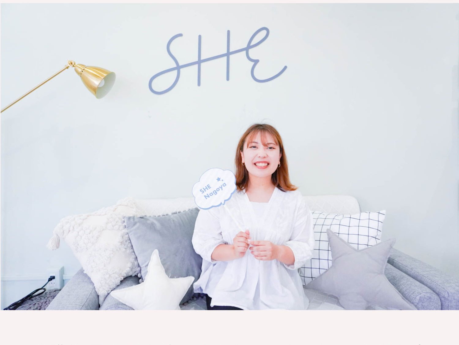 「SHE」ロゴの壁アートを背景に、SHE Nagoyaのうちわを持ちソファに座って微笑むひがめぐさん