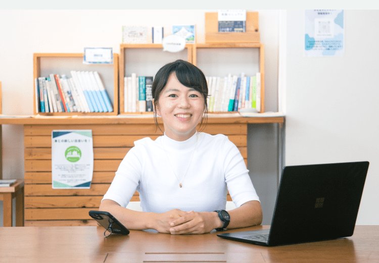 本棚を背に机でノートPCとスマホを前にして微笑むインタビュイー本人