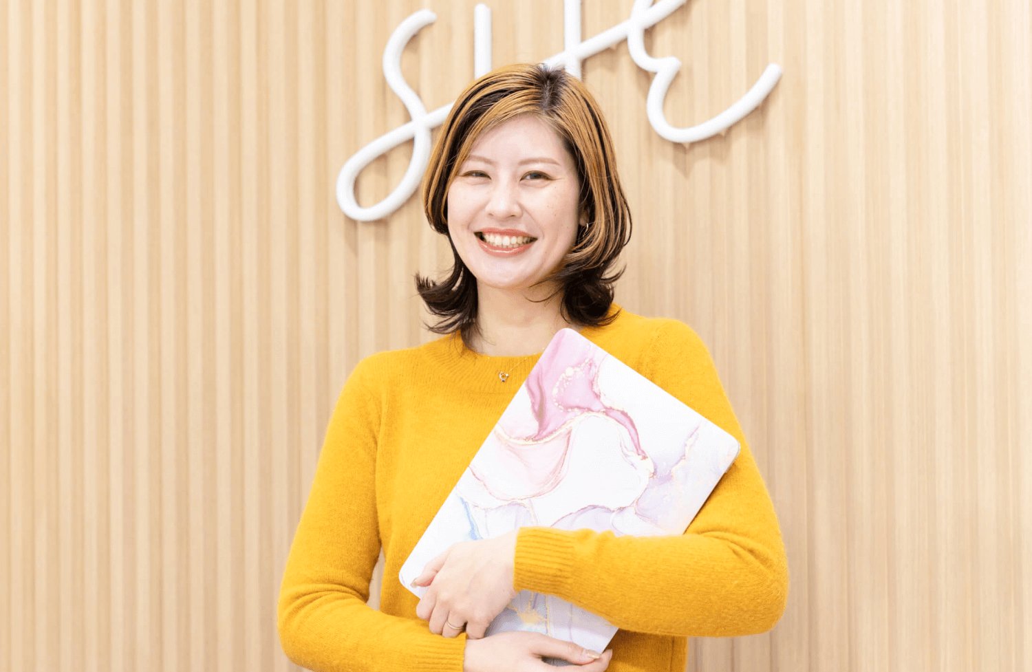 「SHE」ネオンサインの壁の前で大理石柄ノートPCを抱え微笑むhaluさん