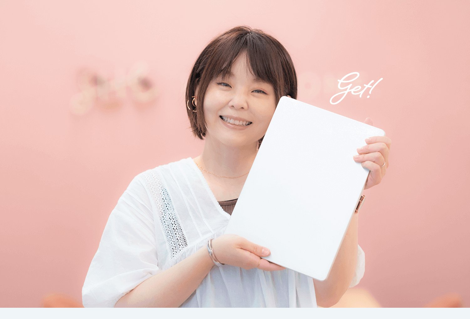 ピンク背景の中、白いノートPCを掲げて笑うのりさん