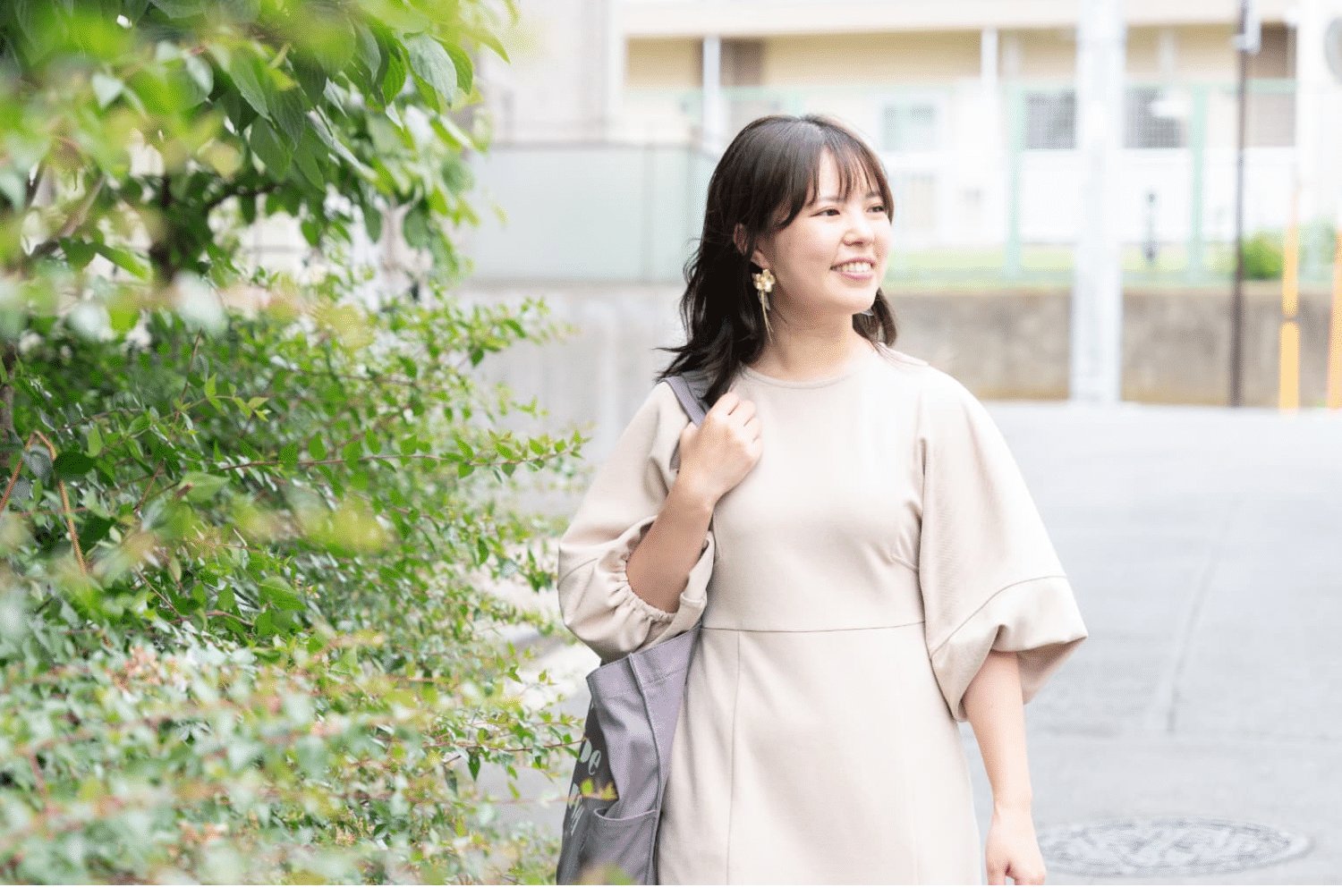 緑の街路樹の脇を歩くベージュのワンピース姿のさやかさん