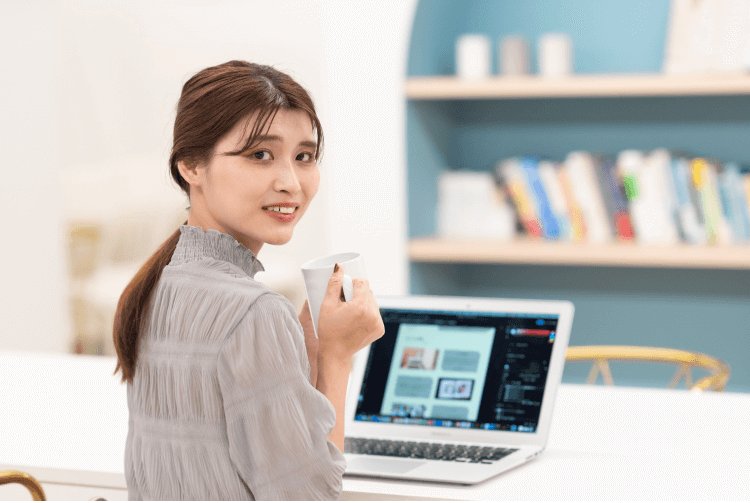 コーヒーカップを持ちながら振り返って微笑む、ノートPCの前に座るさくらさんの写真