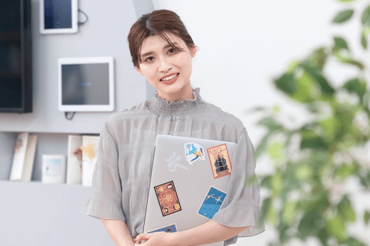 棚と観葉植物のあるオフィスで、ステッカーを貼ったノートPCを抱えて微笑むさくらさんの写真