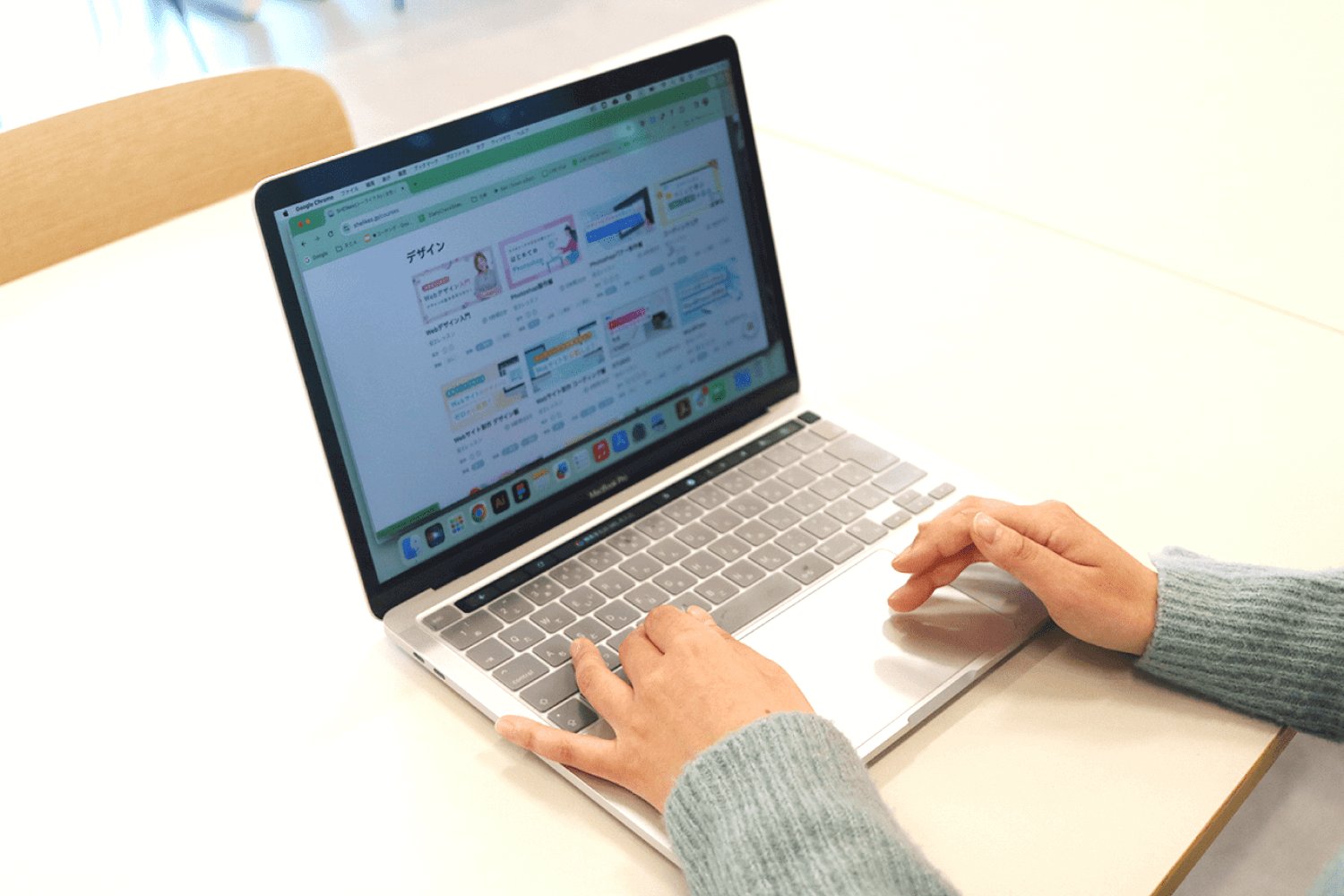 カフェ風の空間のテーブルでノートPCを操作するSAKIさんの手元、画面にはSHElikesのコース一覧