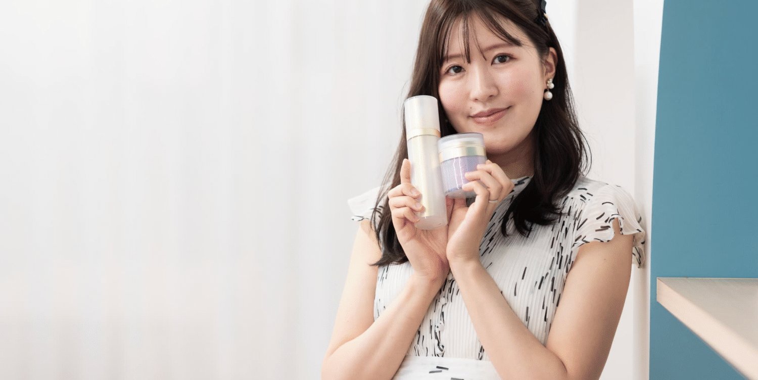 手がけたスキンケア商品(ボトルとジャー)を顔の横に掲げて持つMOEさんのポートレート
