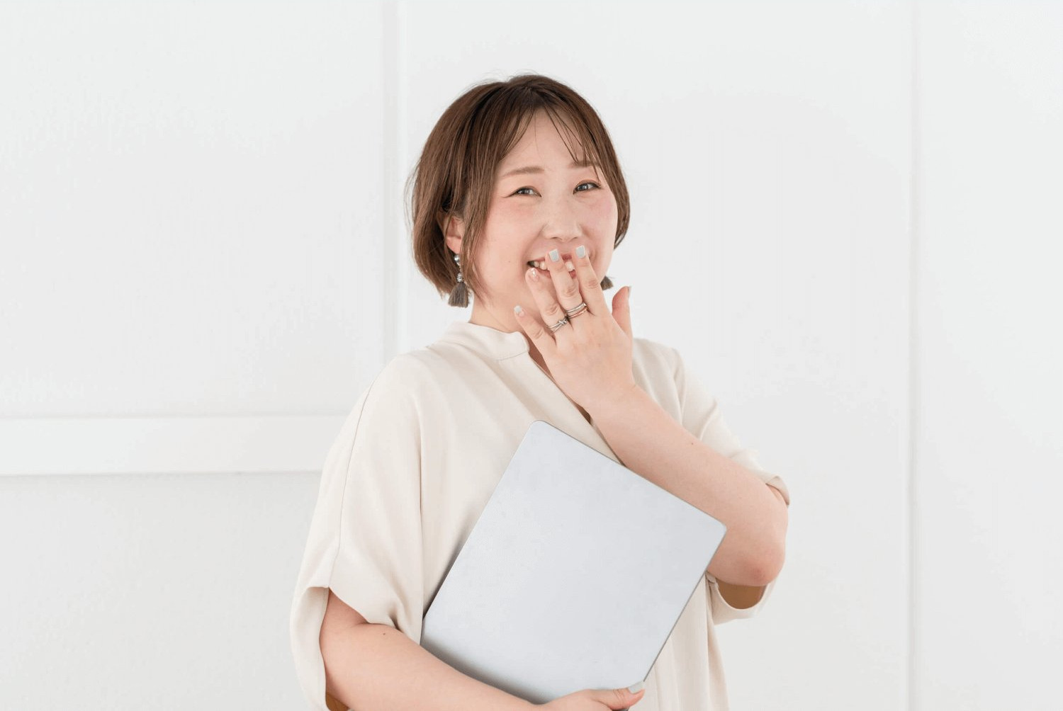 白い壁の前でノートパソコンを抱え、笑顔で口元に手を当てるリエさん