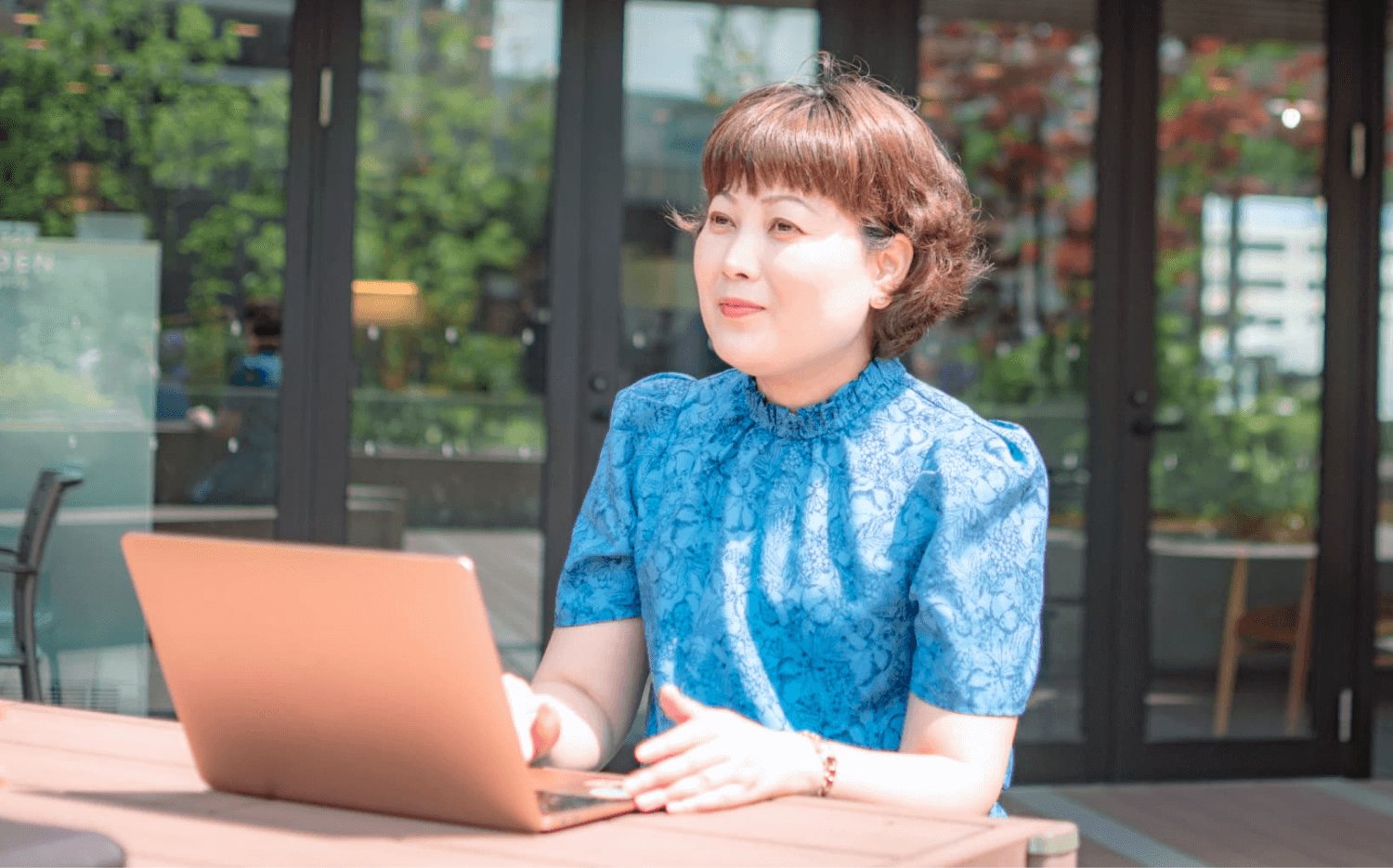 ガラス扉と緑を背景に、屋外のテーブルでノートPCを操作する薫さん