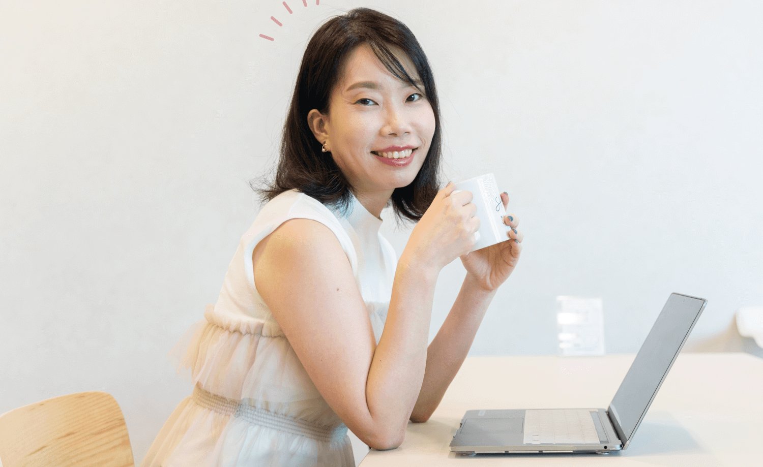 ノートパソコンの前でマグカップを持ち、振り返って微笑む直子さん
