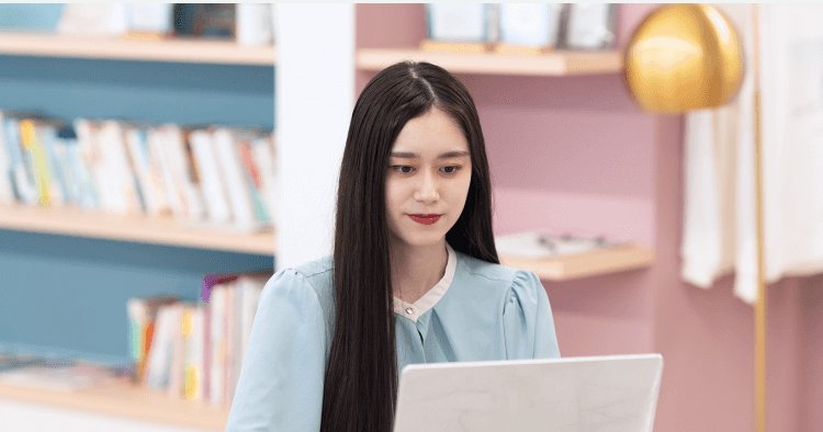 書棚とピンクの壁を背景に、ノートPCを見つめて入力するHinakoさん