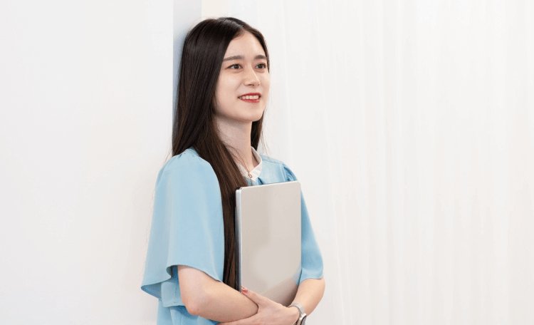 白い壁に寄りかかり、資料フォルダを両手で抱えるHinakoさん