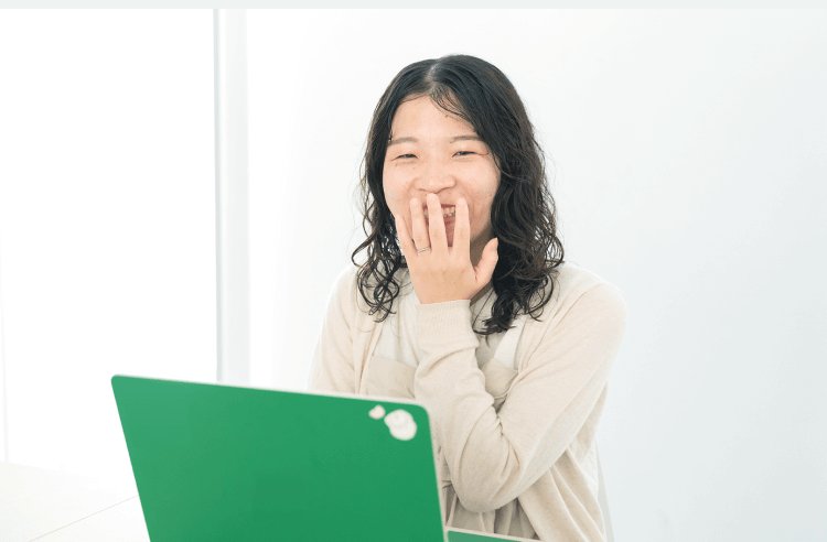 緑のノートPCを操作しながら笑う本人のポートレート。自宅、白い壁を背景に