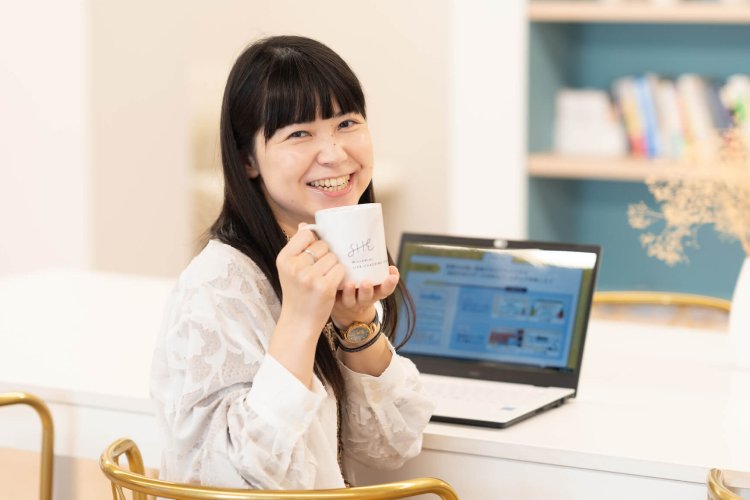 SHEのマグカップを持ち、講座画面を開いたノートPCの前で微笑むちゃみさん