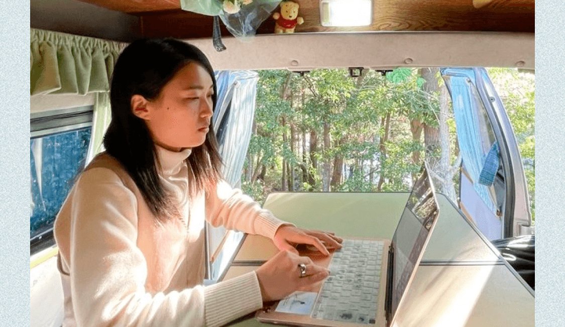 キャンピングカーの車内で、窓の外の木々を背にノートPCで作業する可部ちゃん
