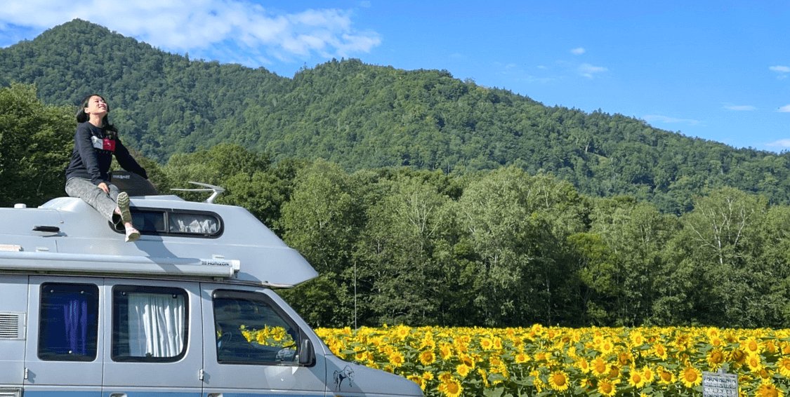 キャンピングカーの屋根に座り空を仰ぐ可部ちゃんと、背景にひまわり畑と山