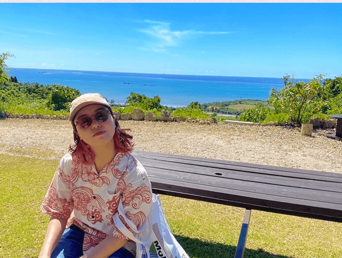 沖縄の海を背景にベンチの前に座る杏珠さん(帽子・サングラス)