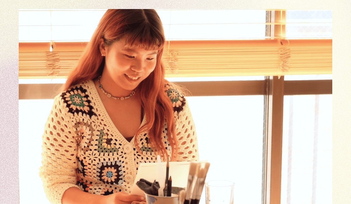 窓際でクロシェのカーディガンを着て、筆を手に微笑むTerakoさん