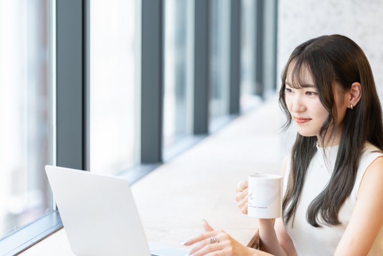 窓際の席でノートPCを開き、マグカップを手に外を見つめるさきさん