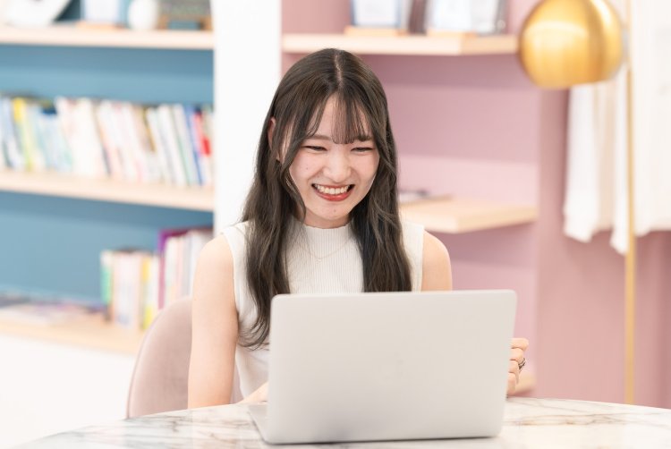 本棚のある会場でノートPCを操作しながら笑顔を見せるさきさん(もくもく会のイメージ)